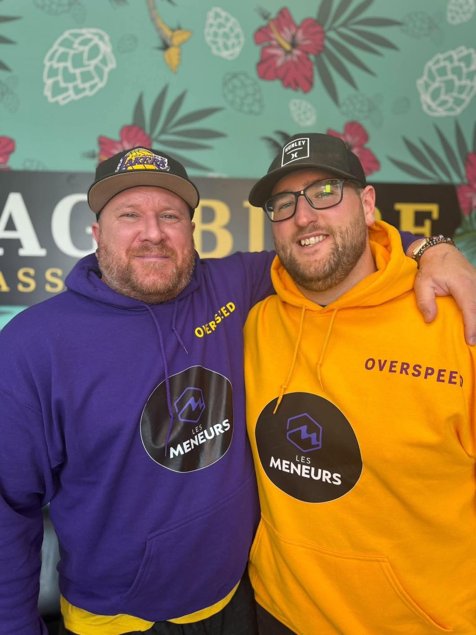 Portrait des deux animateurs des Meneurs Les deux animateurs des Meneurs portant des sweats à capuche violet et jaune devant une fresque florale turquoise.